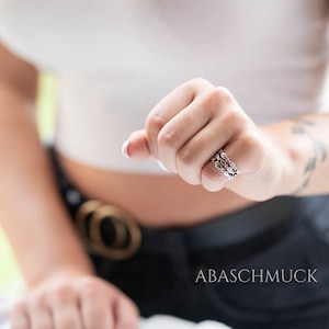 May include: A silver ring with a three-band design, featuring a repeating pattern of leaf-like shapes. The ring is worn on a hand, with the brand name "ABASCHMUCK" visible in the image.