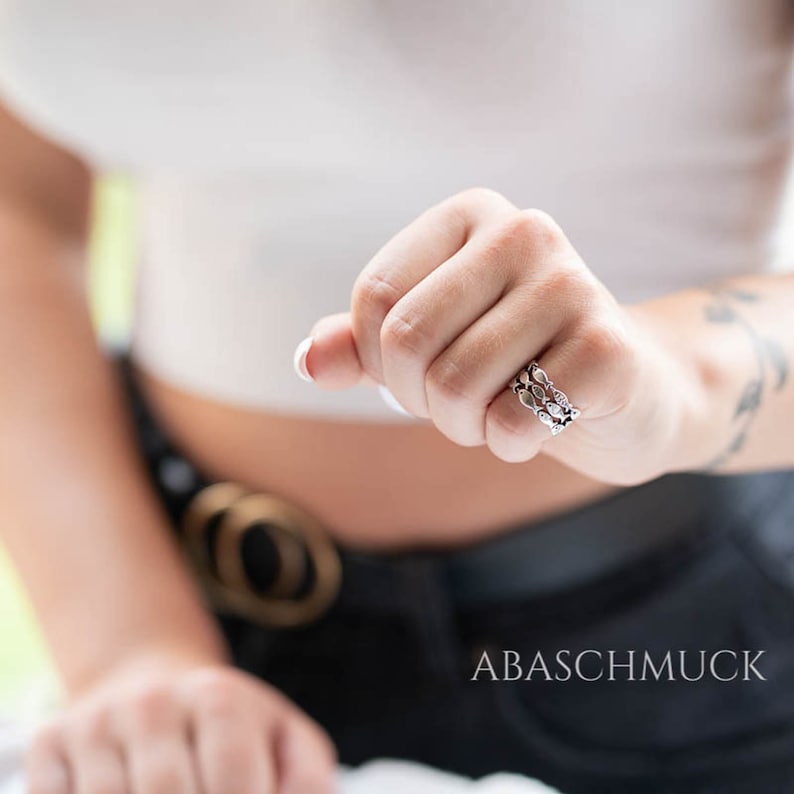 May include: A silver ring with a three-band design, featuring a repeating pattern of leaf-like shapes. The ring is worn on a hand, with the brand name "ABASCHMUCK" visible in the image.