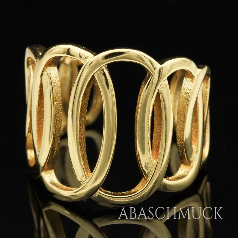 May include: A gold-colored ring with an interwoven design of oval shapes. The ring has a polished finish and is set against a black background. The brand name "ABASCHMUCK" is visible at the bottom.