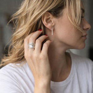 May include: A silver ring with a woven design and a pair of silver bar earrings. The jewelry is worn by a person wearing a white t-shirt. The person has blonde hair and red nail polish.