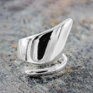 Può includere: Un anello in argento lucido con un design asimmetrico unico. L'anello presenta un elemento curvo e appuntito che si eleva sopra la fascia, creando un aspetto moderno e scultoreo. La fascia si avvolge a spirale, aggiungendo allo stile distintivo dell'anello.