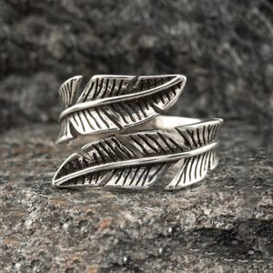 Op de afbeelding: Een zilverkleurige ring met een veermotief. De ring heeft twee overlappende veermotieven, met gedetailleerde adering. De ring ligt op een getextureerd, grijs stenen oppervlak.