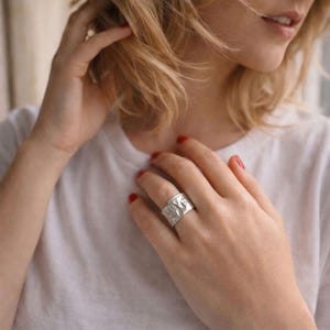 Op de afbeelding: Een brede, zilveren ring met een getextureerd oppervlak wordt aan een vinger gedragen. De ring heeft een gedetailleerd ontwerp, mogelijk een natuurlijke scène of abstract patroon. De ring is het middelpunt en toont een sieraad.