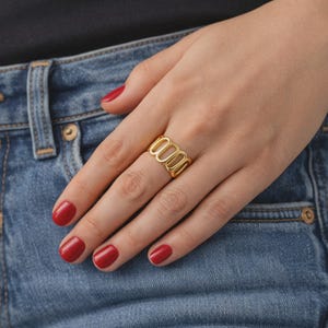 May include: A gold-colored ring with an open, oval design. The ring is worn on a finger, with red nail polish on the nails. The background includes blue denim jeans.