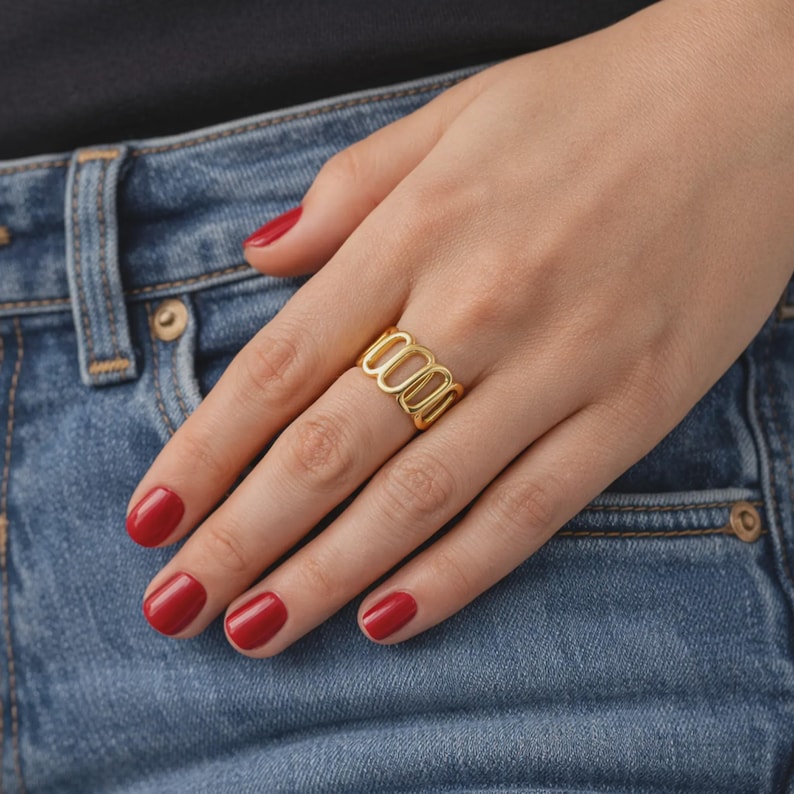 May include: A gold-colored ring with an open, oval design. The ring is worn on a finger, with red nail polish on the nails. The background includes blue denim jeans.