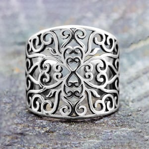May include: A wide, silver-colored ring with an intricate openwork design. The ring features a repeating pattern of swirling lines and heart-shaped elements. The ring is displayed on a textured, gray surface.