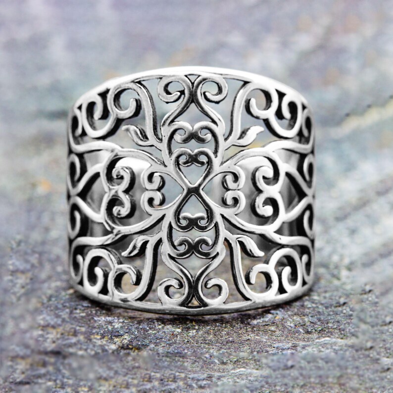 May include: A wide, silver-colored ring with an intricate openwork design. The ring features a repeating pattern of swirling lines and heart-shaped elements. The ring is displayed on a textured, gray surface.