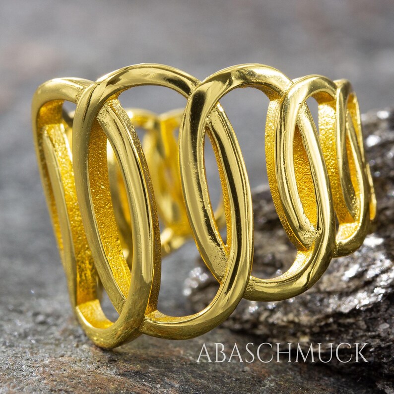 May include: A gold-colored ring with a unique design featuring interconnected oval shapes. The ring is displayed on a textured, gray surface. The brand name "ABASCHMUCK" is visible in the lower right corner.