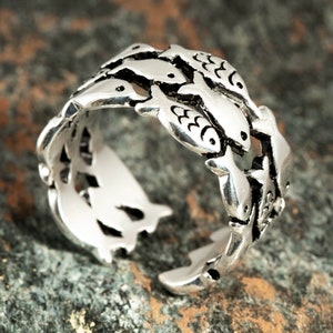 May include: A silver-colored ring featuring a design of small fish swimming around the band. The ring has an open design, allowing for some adjustment in size. The fish are detailed with etched features, creating a unique aquatic theme.