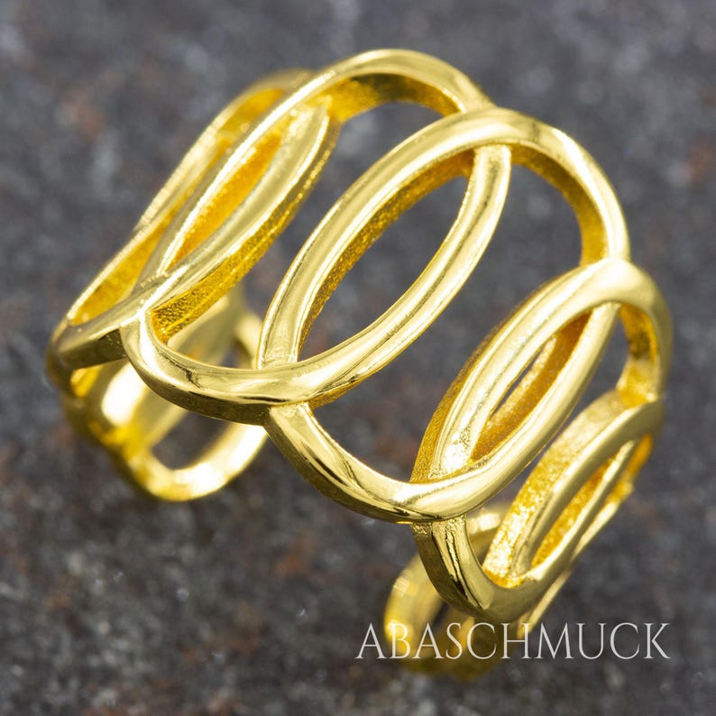 May include: A gold-colored ring with an open, interwoven design. The ring features multiple oval-shaped links that create a continuous, flowing pattern. The ring is made of a shiny, reflective metal and is displayed against a dark background. The text "ABASCHMUCK" is visible.