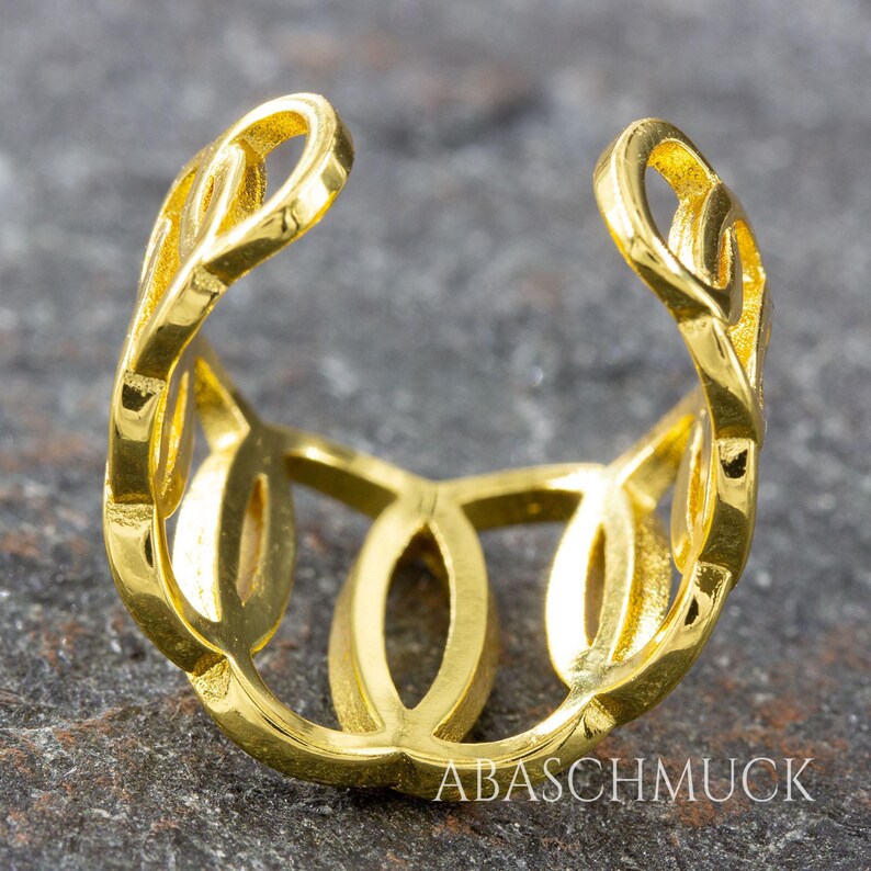 May include: A close-up of a gold-colored ring with an open design. The ring features a series of interconnected oval and leaf-like shapes. The ring is set against a gray, textured surface. The text "ABASCHMUCK" is visible on the ring.