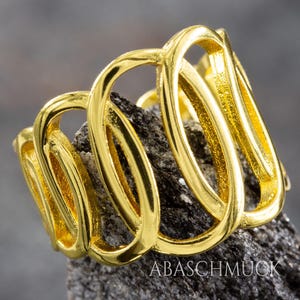 May include: A gold-colored ring with an abstract design featuring interconnected loops. The ring is displayed on a dark, textured surface. The text "ABASCHMUCK" is visible on the surface.