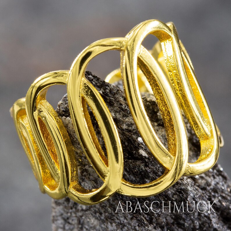 May include: A gold-colored ring with an abstract design featuring interconnected loops. The ring is displayed on a dark, textured surface. The text "ABASCHMUCK" is visible on the surface.