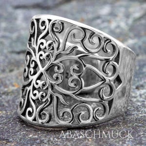 May include: A wide, silver-colored ring with an intricate openwork design. The ring features a detailed pattern of swirling lines and floral motifs. The band is thick and has a textured surface. The text "ABASCHMUCK" is visible on the ring.