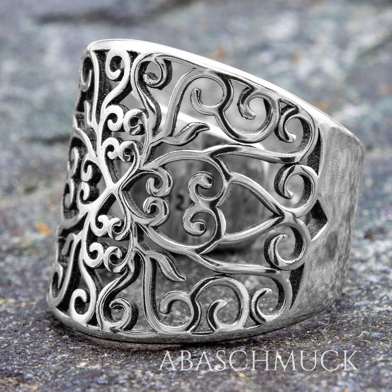 May include: A wide, silver-colored ring with an intricate openwork design. The ring features a detailed pattern of swirling lines and floral motifs. The band is thick and has a textured surface. The text "ABASCHMUCK" is visible on the ring.