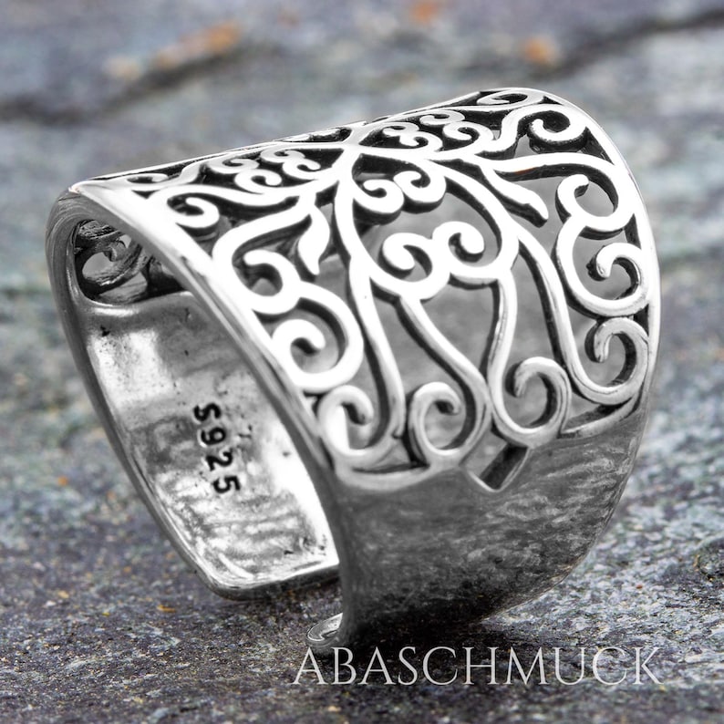 May include: A silver ring with an openwork design. The ring features an intricate pattern of swirling lines and curves. The band is wide and textured, with the text "S925" visible. The brand name "ABASCHMUCK" is also visible.