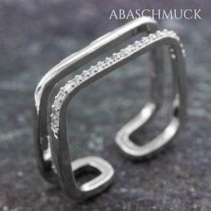May include: Silver ring with a double square design. The outer square is lined with small clear stones.