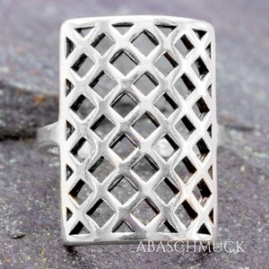May include: A silver ring with a rectangular, openwork design. The ring features a lattice pattern of diamond shapes, creating an airy and geometric aesthetic. The ring is set against a textured, grey surface.