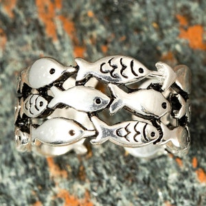 May include: Silver-toned ring featuring a repeating pattern of fish. Each fish has a small, black eye and detailed fins. The ring's design is open, allowing light to pass through the spaces between the fish.