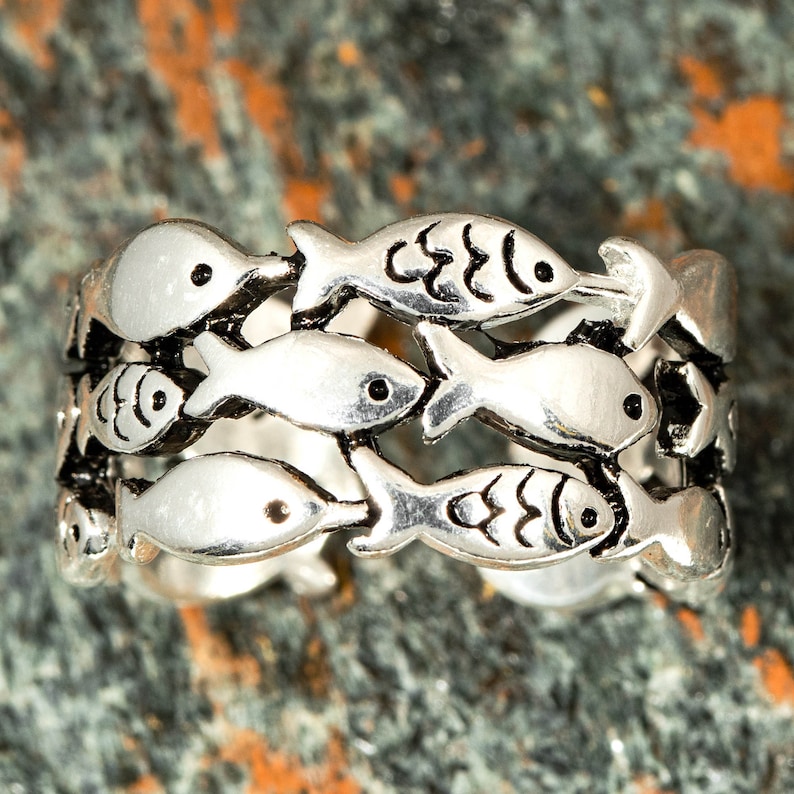 May include: Silver-toned ring featuring a repeating pattern of fish. Each fish has a small, black eye and detailed fins. The ring's design is open, allowing light to pass through the spaces between the fish.