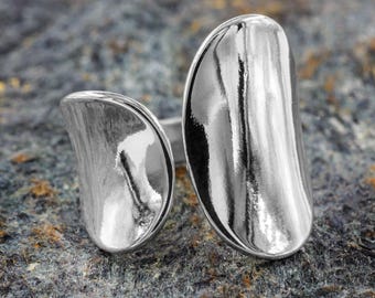 Puede incluir: Un anillo plateado brillante con dos grandes formas oblongas curvas. El anillo tiene un diseño moderno y minimalista. El metal pulido refleja la luz, creando una apariencia brillante y llamativa. El anillo está sobre una superficie texturizada gris y marrón.