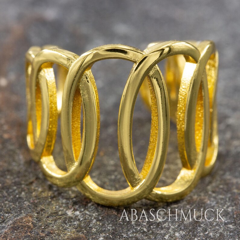 May include: A gold-colored ring with an open, interwoven design. The ring features a series of connected oval shapes, creating a continuous loop pattern. The ring is made of a shiny, reflective metal, and the brand name "ABASCHMUCK" is visible.