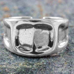 Peut inclure: Gros plan d'une bague argentée avec un motif géométrique texturé. La bague a un large anneau avec une série de formes rectangulaires. La bague est posée sur une surface mouchetée gris-vert.