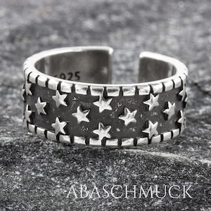 Könnte beinhalten: Ein verstellbarer Silberring mit einem schwarzen Band und einem sich wiederholenden Muster aus silbernen Sternen. Der Ring ist mit der Nummer "925" gekennzeichnet.