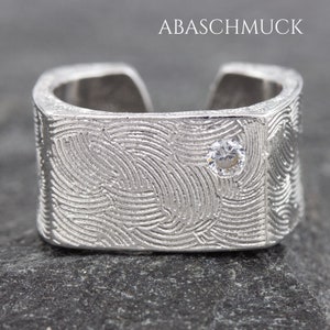 Puede incluir: Un anillo de plata con un diseño texturizado y una pequeña piedra transparente. El anillo es ancho y tiene una forma cuadrada.