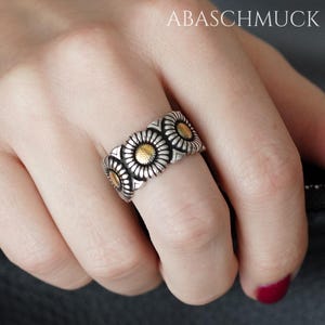 Op de afbeelding: Een zilveren ring met een herhalend madeliefjesontwerp. Elke madelief heeft een goudkleurig midden en gedetailleerde zilveren bloemblaadjes. De ring wordt aan een vinger gedragen, met de merknaam "ABASCHMUCK" zichtbaar.