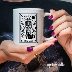 May include: White ceramic mug with a black and white illustration of a tarot card depicting a skeleton holding a cat. The card reads "The Cat".