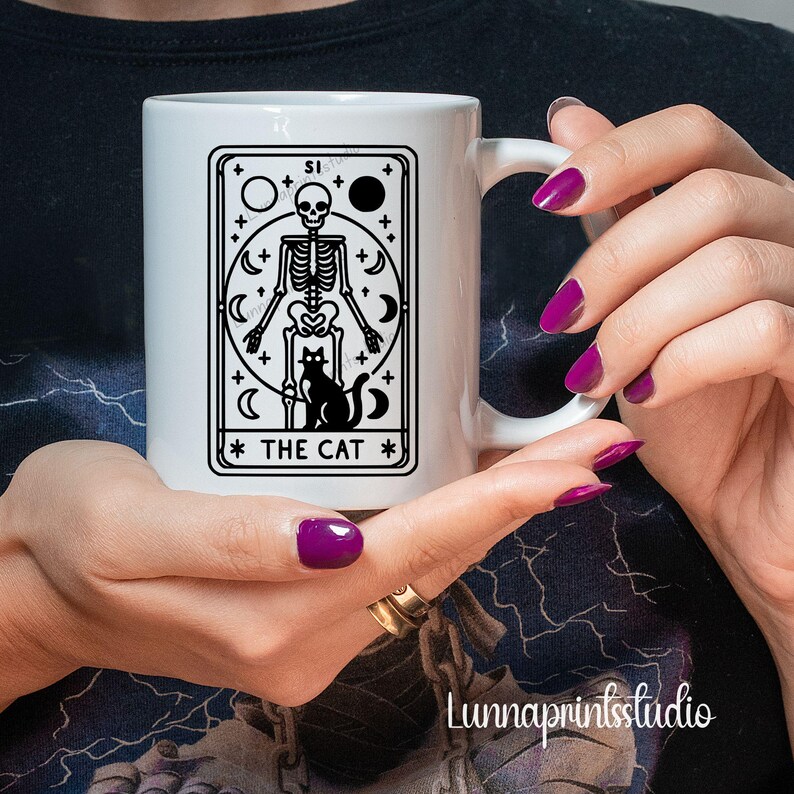 May include: White ceramic mug with a black and white illustration of a tarot card depicting a skeleton holding a cat. The card reads "The Cat".