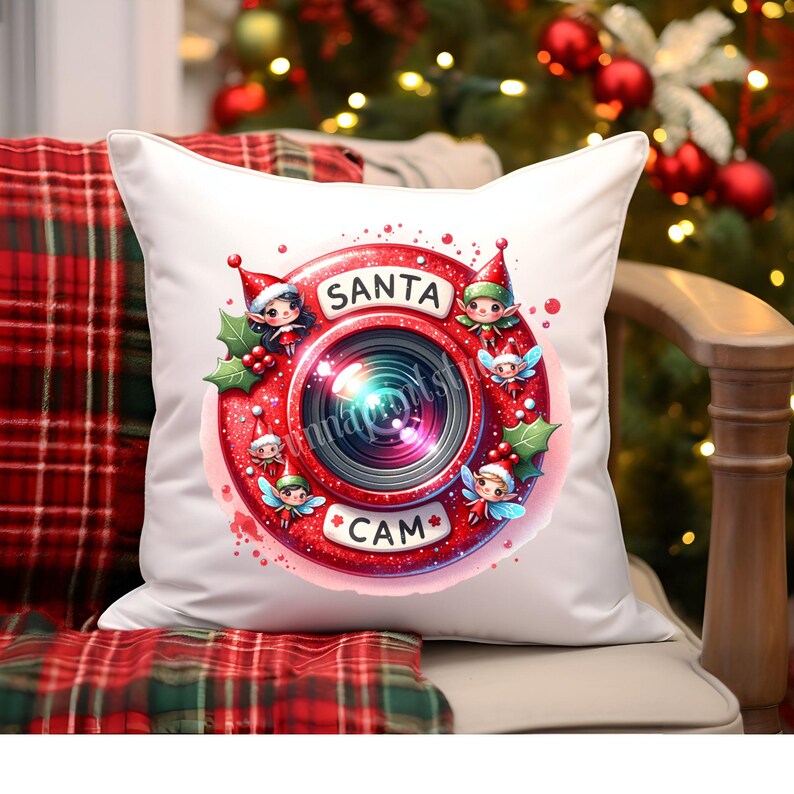 Puede incluir: Almohada blanca con un dise&ntilde;o de Santa Cam rojo y blanco. El dise&ntilde;o presenta un objetivo de c&aacute;mara rojo con un duende verde y blanco asom&aacute;ndose por el objetivo. El texto "Santa Cam" est&aacute; escrito en la c&aacute;mara.