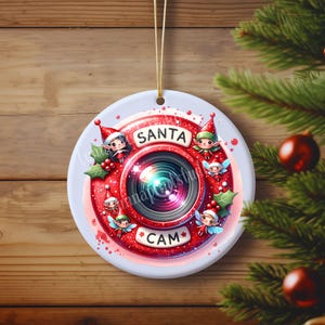 Puede incluir: Un adorno navide&ntilde;o rojo y blanco con una lente de c&aacute;mara con las palabras "Santa Cam" y rodeado de duendes.