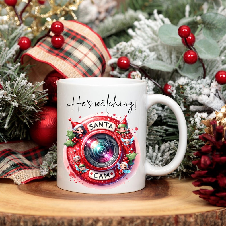 Puede incluir: Taza de cer&aacute;mica blanca con una ilustraci&oacute;n roja y blanca de una c&aacute;mara de Santa Claus rodeada de duendes. El texto "He's watching!" est&aacute; por encima de la c&aacute;mara y "Santa Cam" est&aacute; por debajo de la c&aacute;mara.