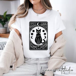 May include: White t-shirt with a black and white graphic of a black cat with a third eye and a crescent moon. The graphic is surrounded by a circle of moons in different phases. The text "Moon: The Black Cat" is below the graphic.