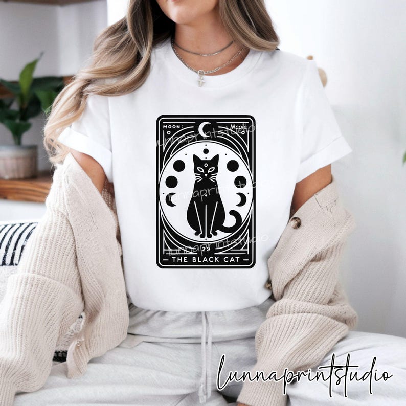 May include: White t-shirt with a black and white graphic of a black cat with a third eye and a crescent moon. The graphic is surrounded by a circle of moons in different phases. The text "Moon: The Black Cat" is below the graphic.