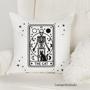 May include: White throw pillow with a black and white illustration of a tarot card depicting a skeleton holding a cat. The card reads "The Cat" and is surrounded by stars.