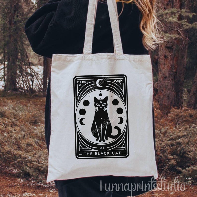 May include: White canvas tote bag with a black and white illustration of a black cat with a third eye, surrounded by moons. The text "Moon Magic" and "The Black Cat" are printed on the bag.
