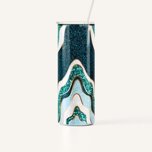 May include: A teal glitter tumbler with a wavy design in shades of teal, white, and light blue, outlined in gold. The tumbler has a silver lid and a silver straw. The design resembles an abstract landscape.