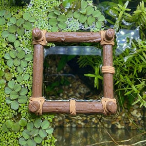 May include: A brown, square aquarium decoration resembling a rustic wooden frame, with rope accents at the corners. The frame is partially submerged in water, surrounded by aquatic plants and greenery, creating a naturalistic scene.