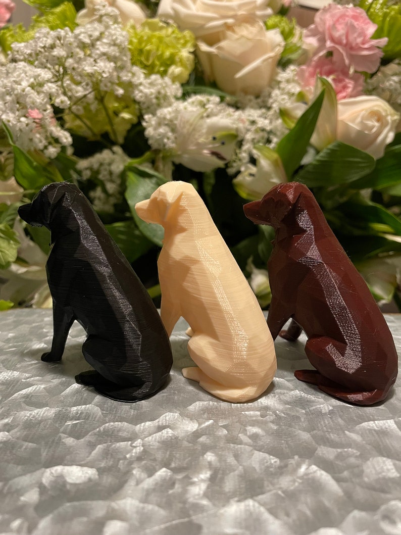 K&ouml;nnte beinhalten: Drei 3D-gedruckte Hundefiguren in Schwarz, Wei&szlig; und Braun, die auf einer strukturierten Oberfl&auml;che sitzen.