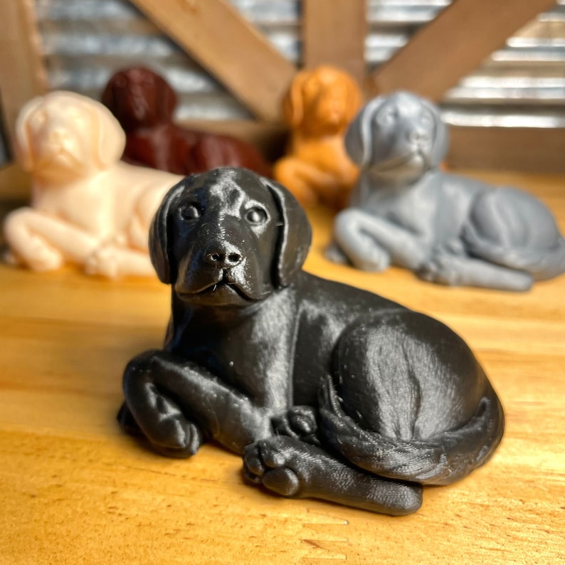 May include: A collection of five small, 3D printed dog figurines. The figurines are in various colors, including black, white, brown, and gray. The figurines are all in a sitting position with their heads tilted slightly upwards.