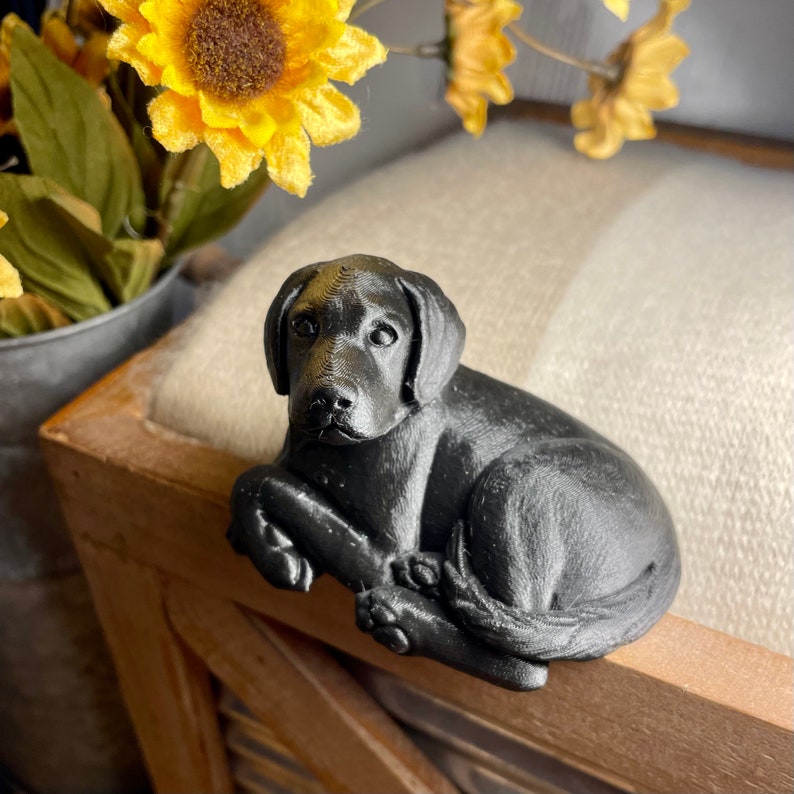 May include: A black Labrador Retriever dog figurine, sitting with its paws tucked under its body. The figurine is made of a smooth, black material and has a realistic appearance.