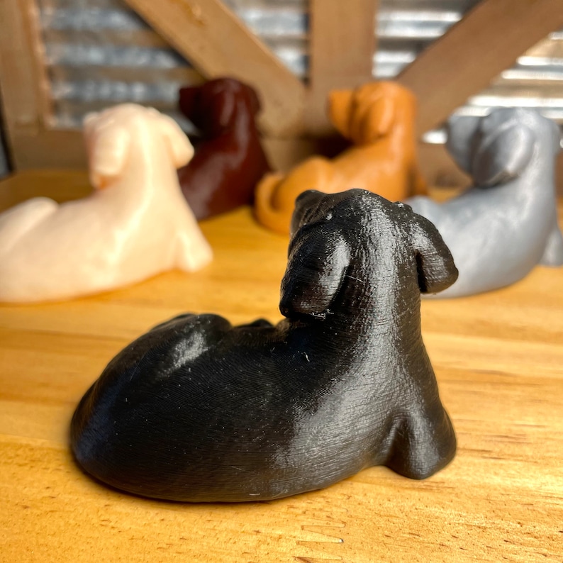 May include: A collection of five small, 3D printed dachshund figurines in various colors, including black, brown, white, and gray. The figurines are posed in a sitting or lying down position.