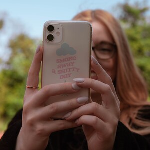 May include: A clear phone case with a pink and white design. The design features a cloud with the text "Smoke away a shitty day".