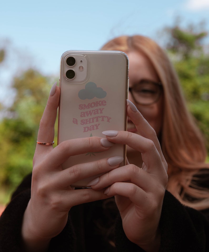 May include: A clear phone case with a pink and white design. The design features a cloud with the text "Smoke away a shitty day".