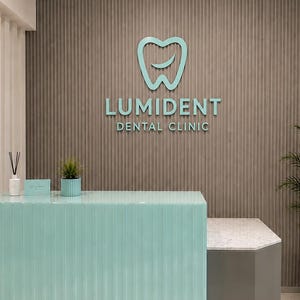 May include: A reception desk with a light mint green fluted front and a white marble top. On the wall behind, a light mint green logo of a tooth with a smile and the text LUMIDENT DENTAL CLINIC is mounted on a brown vertical slatted wall.