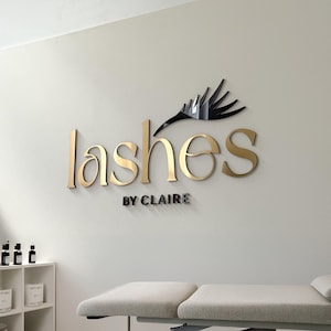 Op de afbeelding: Een bord aan de muur voor een wimpersalon. Het woord "lashes" staat in gouden, gestileerde letters, met "BY CLAIRE" in kleinere, grijze letters eronder. Een zwarte wimpergrafiek staat boven het woord "lashes".