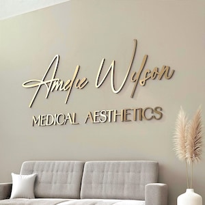 May include: Gold-coloured sign on a beige wall. The sign reads "Amelie Wilson" in cursive script above the words "MEDICAL AESTHETICS" in block letters. A grey sofa and a vase with decorative plants are in the foreground.